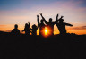 A group of adventurous hikers with arms outstretched shown silhouetted against a red sunset.